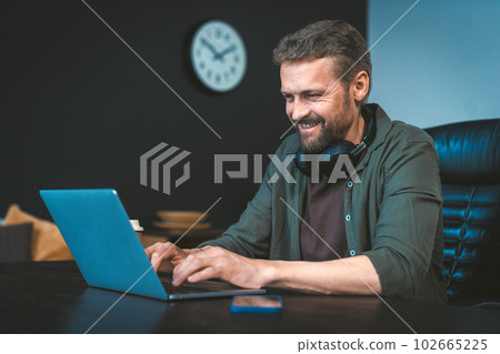 Happy and motivated man is working on laptop at desk in stylish loft office, with intention of making money. He focused and determined, yet also relaxed and comfortable in work environment. High 102665225
