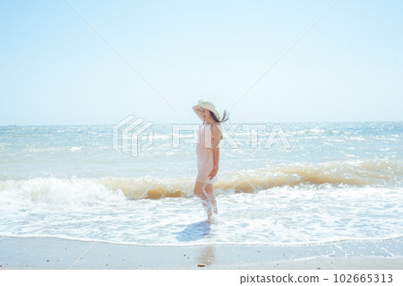 A woman in a pink dress and straw hat walking on the beach 102665313