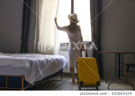 Portrait of tourist woman standing nearly window, looking to beautiful view with her luggage in hotel bedroom after check-in. 102665424