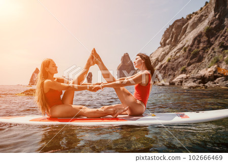 Woman sup yoga. Two happy sporty women practising yoga pilates on paddle sup surfboard. Female stretching doing workout on sea water. Modern individual female hipster outdoor summer sport activity. 102666469