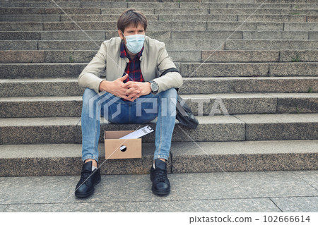 Sad businessman in a medical mask sitting on the stairs outdoors with a box of documents. 102666614