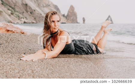 Woman summer travel sea. Happy tourist in black dress enjoy taking picture outdoors for memories. Woman traveler posing on sea beach surrounded by volcanic mountains, sharing travel adventure journey 102666639