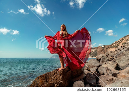 Red dress sea woman. A blonde with flowing hair in a long flowing red dress stands on a rock near the sea. Travel concept, photo session at sea Red dress sea woman. A blonde with flowing hair in a long flowing red dress stands on a rock near the sea. Travel concept, photo session at sea 102668165