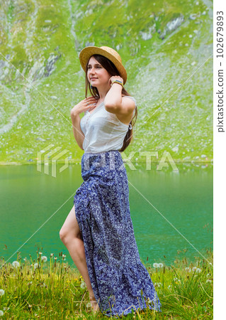 A beautiful woman in a dress and hat stands near of Lake Balea A beautiful woman in a dress and hat stands near of Lake Balea 102679893