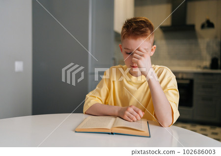 Portrait of focused cute little student boy reading paper book following lines with finger along page sitting at table in living room. Redhaired schoolboy learning at home with textbook. 102686003