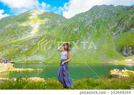 A beautiful woman in a dress and hat stands near of Lake Balea A beautiful woman in a dress and hat stands near of Lake Balea 102689269