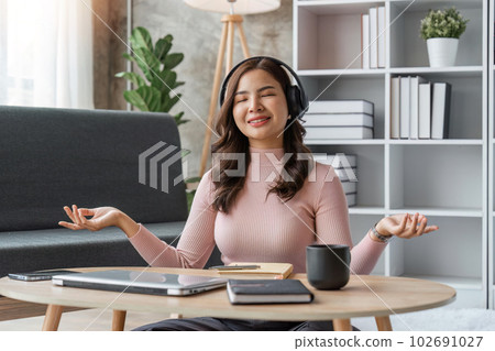 woman listening to music, using headphones, sitting on couch in living room, Happy young lady with closed eyes enjoying music 102691027