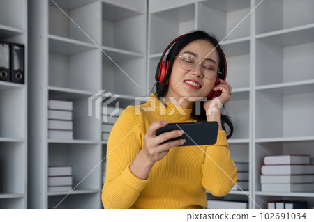 woman in with mobile phone listening music in headphones and relax at home. Happy girl with closed eye sitting on couch in living room 102691034