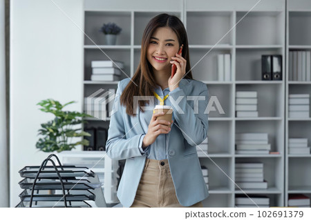 Confident business woman attractive smiling holding coffee talking on the phone and looking at camera in smart in office Confident business woman attractive smiling holding coffee talking on the phone and looking at camera in smart in office 102691379