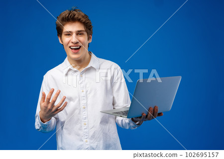 Young man standing in shirt, holding laptop and looking at camera on blue background 102695157