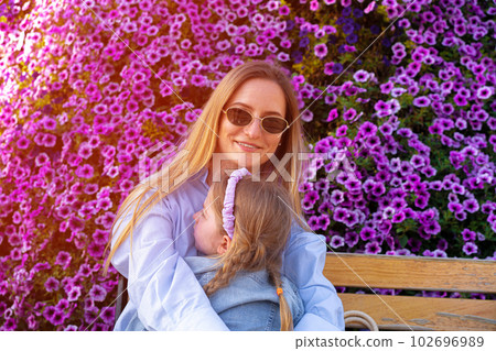 Young woman sitting on the bench with her little daughter near wall of petunia flowers Young woman sitting on the bench with her little daughter near wall of petunia flowers 102696989