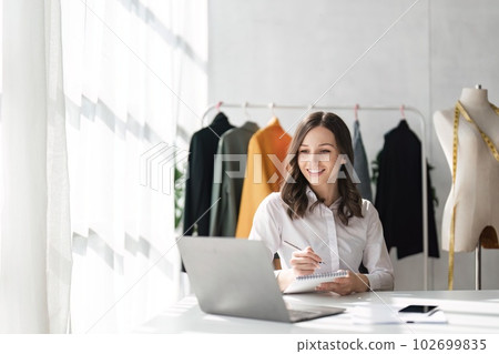 Young attractive female fashion designer sketching idea on paper at desk, working with a laptop at home Young attractive female fashion designer sketching idea on paper at desk, working with a laptop at home 102699835