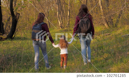Happy family life concept. young women walk through forest with small child with backpacks their backs. walk woods summer weekend sunset. little girl holding hands parents walking along path summer Happy family life concept. young women walk through forest with small child with backpacks their backs. walk woods summer weekend sunset. little girl holding hands parents walking along path summer 102701285