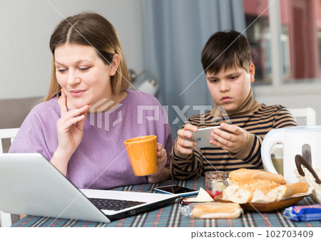 Young woman working at laptop and son using phone at table with tea Young woman working at laptop and son using phone at table with tea 102703409