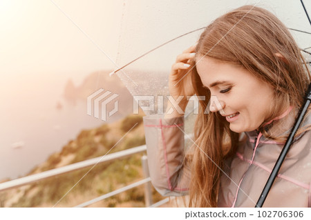 Woman rain umbrella. Happy woman portrait wearing a raincoat with transparent umbrella outdoors on rainy day in park near sea. Girl on the nature on rainy overcast day. 102706306