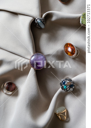 Many different silver rings with various natural gemstones on a beige background. 102713371