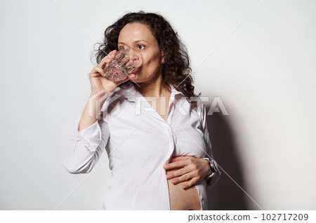Beautiful adult Latin American pregnant woman, touching her bare belly, taking dieting nutritional supplement with vitamins minerals and drinking water, isolated on white background. Healthy pregnancy 102717209