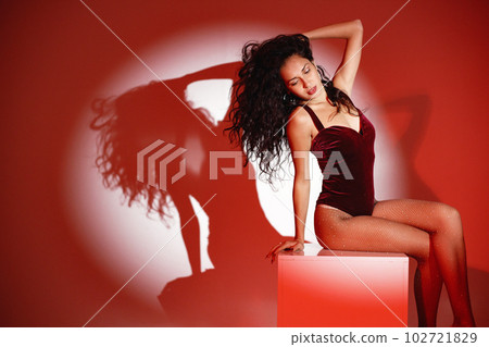 Curly brunette girl sitting on a white cube and posing for a photo. Girl wearing velour bodysuit. Projectors light girl up. 102721829