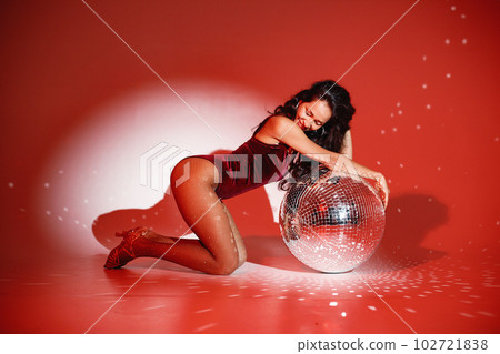 Curly brunette girl sitting near big disco ball and posing for a photo. Girl wearing velour bodysuit. Projectors light girl up. Curly brunette girl sitting near big disco ball and posing for a photo. Girl wearing velour bodysuit. Projectors light girl up. 102721838