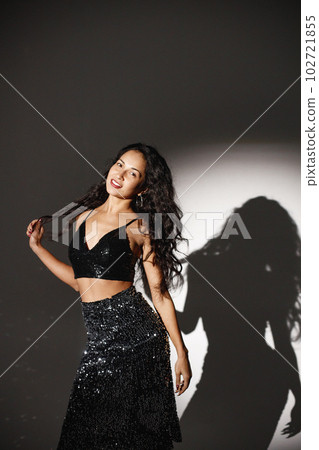 Curly brunette girl standing near white wall and posing for a photo. Girl wearing black skirt and top. Projectors light girl up. 102721855