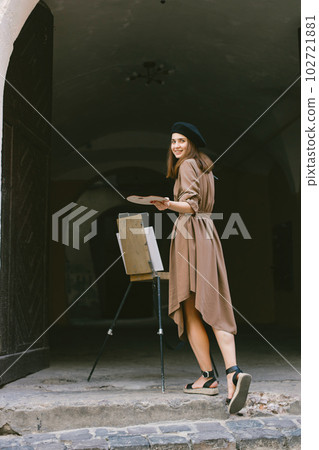 Female painter painting in the city. A woman wearing khaki dress and a black hat. Girl drawing a picture on an easel in summer on a street. 102721881