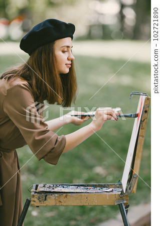 Female painter painting in the park. A woman wearing khaki dress and a black hat. Girl drawing a picture on an easel in summer on nature. 102721890