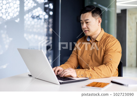 Serious and focused businessman working with laptop, man in glasses and shirt inside office building, asian boss typing on computer. Serious and focused businessman working with laptop, man in glasses and shirt inside office building, asian boss typing on computer. 102723090
