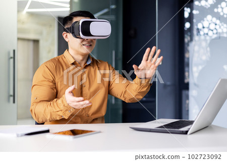 Successful businessman in online meeting uses vr glasses virtual reality, man communicates remotely with partners in metaverse, works inside office sitting at workplace with laptop. 102723092