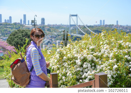 Young beautiful woman tourist with a backpack looks at the camera on the background of the urban landscape overlooking the Fatih Sultan Mehmet Bridge. Back view. Otagtepe Park. Young beautiful woman tourist with a backpack looks at the camera on the background of the urban landscape overlooking the Fatih Sultan Mehmet Bridge. Back view. Otagtepe Park. 102726011