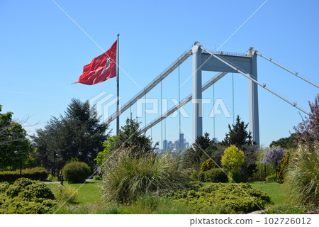 The Turkish flag is waving in the wind in a park overlooking the bridge over the Bosphorus and beautifully blooming sakura. Travel Istanbul background photo. 102726012