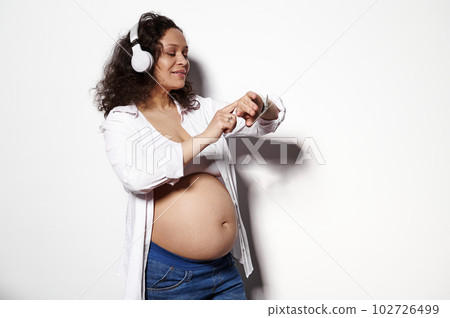 Smiling pregnant woman with big belly advanced pregnancy, wearing wireless headphone, listening to music, adjusting cool soundtrack on her smart wristwatch, isolated on white background. Copy space 102726499