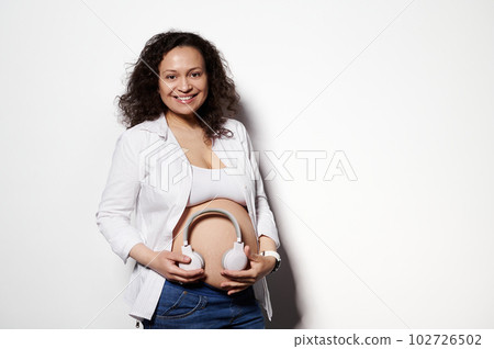 Happy pregnant woman smiling at camera, holding wireless headphones on her bare belly, putting some soothing music for her newborn baby, isolated white background. Maternity. Happy carefree pregnancy Happy pregnant woman smiling at camera, holding wireless headphones on her bare belly, putting some soothing music for her newborn baby, isolated white background. Maternity. Happy carefree pregnancy 102726502