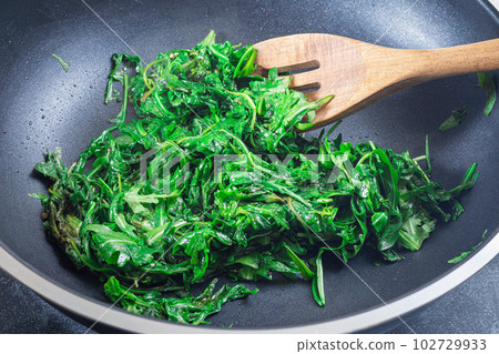 Wilted arugula or rocket salad, preparing a side dish to steak, in frying pan, horizontal 102729933