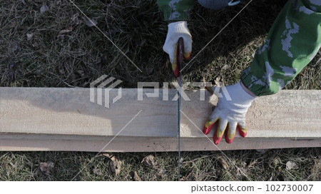 top view of male hands in gloves with a hacksaw sawing a long board on the ground with dry grass, a fragment of construction work with wooden material and a carpenter's hand tool 102730007