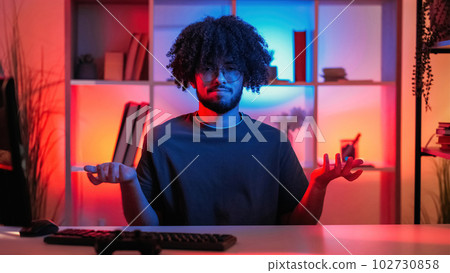 Shrugging gesture. Confused man. Silly expression. Clueless funny guy raising hands sitting desk in dark neon light home interior. Shrugging gesture. Confused man. Silly expression. Clueless funny guy raising hands sitting desk in dark neon light home interior. 102730858