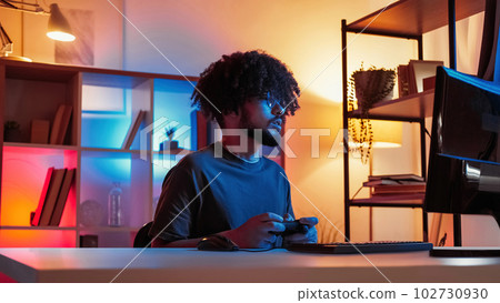 Cyber game. Hipster man. Competition fun. Interested guy playing on computer using console sitting dark neon light home interior. 102730930