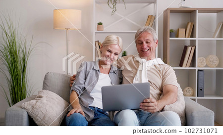 Online conference. Happy parents. Family contacts. Smiling middle-aged man and woman having video call on laptop sitting light home interior. 102731071