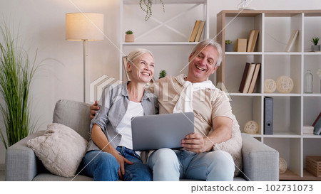 Online connection. Happy couple. Family leisure. Excited middle-aged man and woman sitting sofa with laptop in light home interior. 102731073