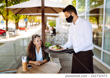Young waiter in protective face mask and gloves while bringing food to a customer in cafe. Covid-19. 102732473