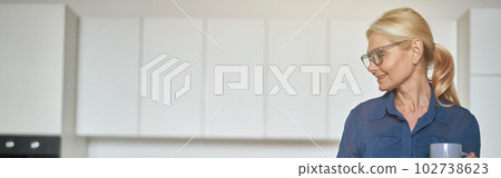 Attractive blonde businesswoman in glasses having a cup of coffee and using laptop in the morning, standing in the kitchen while working from home Attractive blonde businesswoman in glasses having a cup of coffee and using laptop in the morning, standing in the kitchen while working from home 102738623