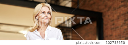 Successful mature blonde business lady looking aside while posing, standing in the office on a daytime Successful mature blonde business lady looking aside while posing, standing in the office on a daytime 102738650