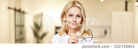 Portrait of confident mature blonde businesswoman looking at camera, holding glasses while posing, standing in the office Portrait of confident mature blonde businesswoman looking at camera, holding glasses while posing, standing in the office 102738661