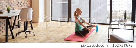 Mature sportswoman sitting in Lotus pose on a mat and using laptop while practicing yoga at home in modern apartment 102738675