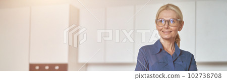Confident mature businesswoman in glasses smiling aside, working at home using laptop, standing with arms crossed in modern kitchen 102738708