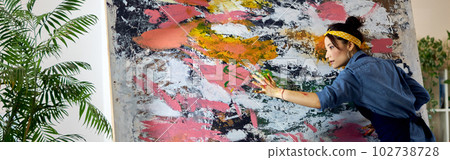 Inspired female artist working on a large modern abstract oil painting, applying paint on canvas with fingers, standing in home studio workshop 102738728