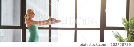 Side view of athletic blonde woman in sportswear exercising using resistance band, following online video tutorial on a laptop, standing on yoga mat at home in front of big window 102738729