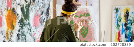 Back view of young female artist in apron painting picture, standing in living room at home Back view of young female artist in apron painting picture, standing in living room at home 102738889