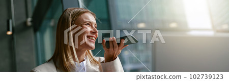 Happy businesswoman recording audio message on phone while while sitting in cozy cafe near windows 102739123