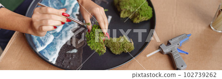 Close up of woman decorator making green ikebana on tray with epoxy resin in florist workshop Close up of woman decorator making green ikebana on tray with epoxy resin in florist workshop 102739262