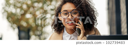 Smiling afro american businesswoman talking phone while working online sitting in cafe terrace 102739483